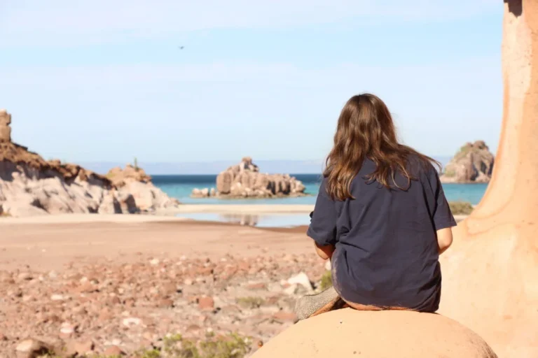 Un momento de calma en tu escape a Espíritu Santo. Vista desde atrás de una persona sentada sobre una roca color terracota, observando la playa desierta y el mar turquesa.