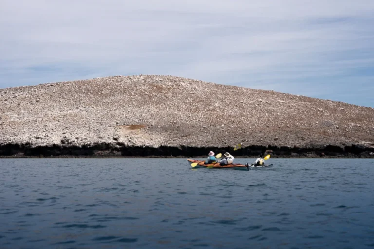 Dos personas reman en un kayak de expedición durante su exploración costera, con un cerro de roca clara dominando el fondo.