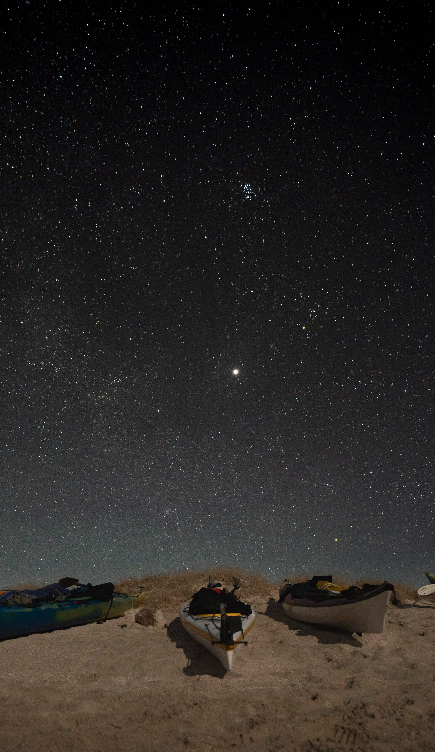 El cielo nocturno lleno de estrellas y la constelación de Pléyades sobre kayaks descansando en la playa de un sitio visitado en los Tours desde Loreto Baja California Sur