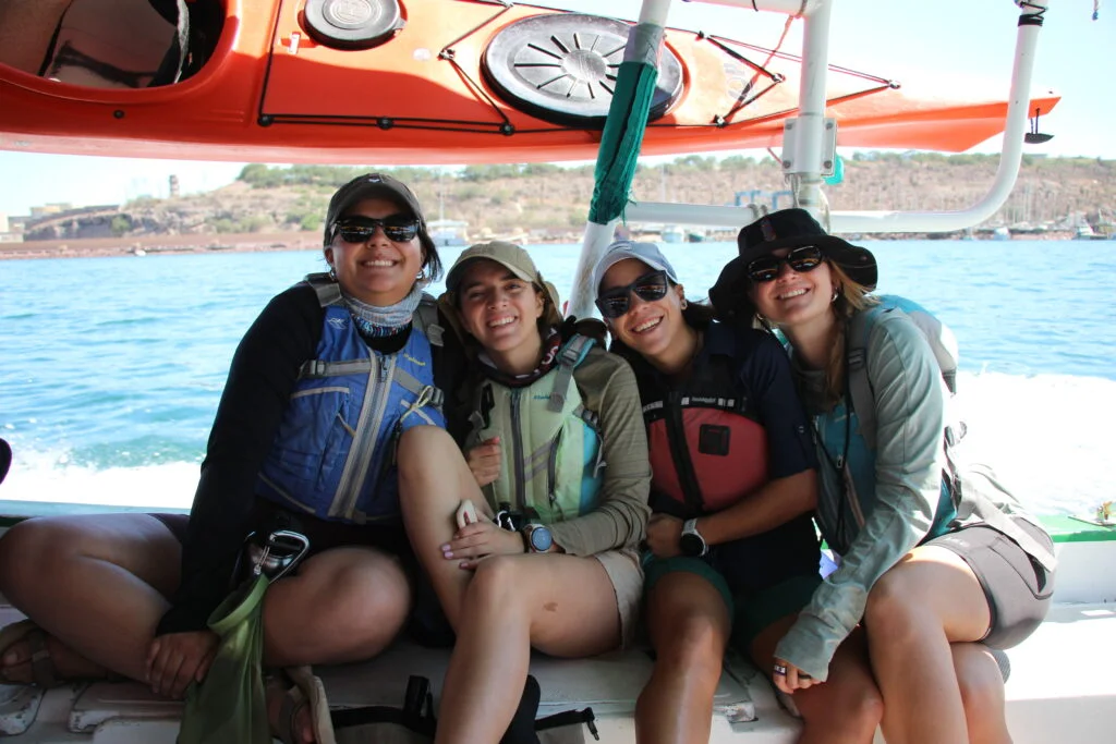 Cuatro mujeres, usando chalecos salvavidas y gorras, posan sonrientes bajo la cubierta de un bote durante sus viajes privados en kayak.